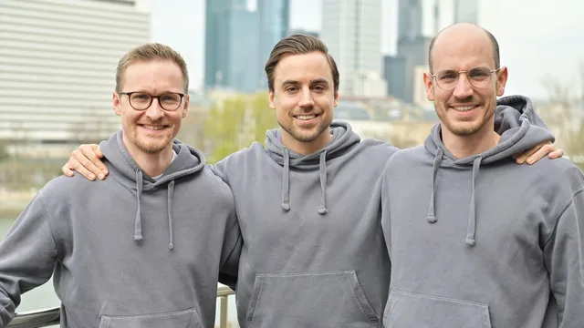 Das Abgebildet ist das Cyreen-Gründerteam (v.l.n.r.): Benedict Boos, Ciril Hofmann und Davide Scheidegger. Alle drei Gründer stehen vor der Frankfurter Skyline und tragen einen grauen Pullover und ein breites Lächeln.