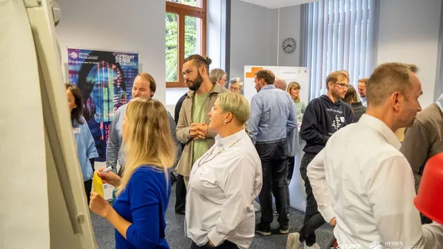 Menschen stehen in einem Raum mit Flipcharts und Postern, einige halten Notizzettel, Fenster mit Lamellenvorhängen im Hintergrund.