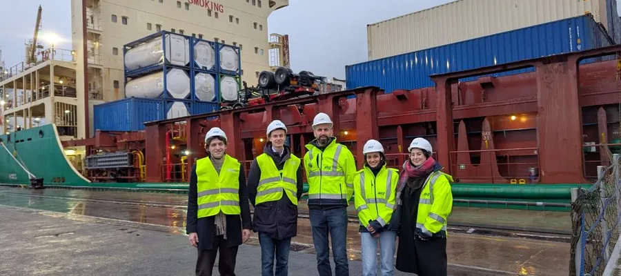 Fünf Personen mit Schutzhelmen und Warnwesten stehen vor einem Frachtschiff mit Containern im Hafen