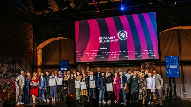 Gruppe von Menschen auf Bühne bei Veranstaltung mit großer Leinwand im Hintergrund, auf der 'Hessischer Gründerpreis Marburg' steht.