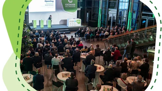 Publikum sitzt an runden Tischen in einem großen Saal und hört einer Person auf einer Bühne mit Präsentation zu.