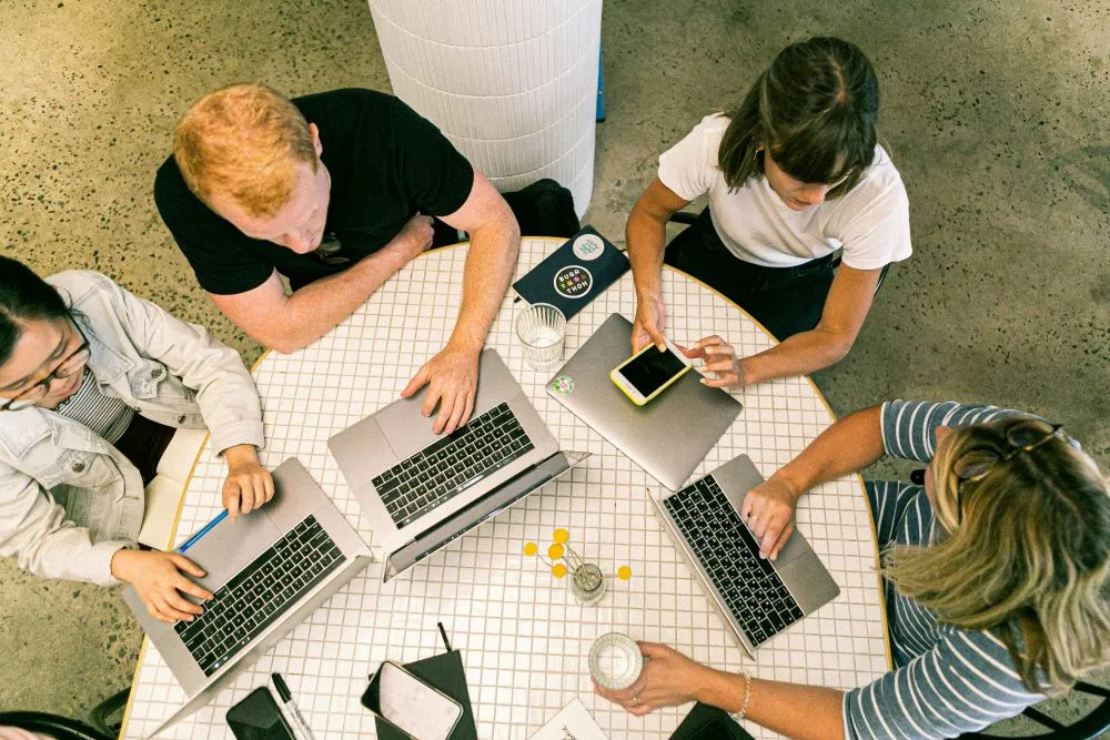 Vier junge Menschen sitzen an einem runden Tisch. Auf diesem Tisch stehen Laptops, Handy. Die dargestellten Menschen arbeiten zusammen.